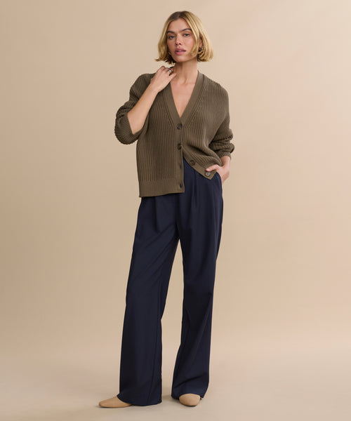 A woman with short blonde hair poses confidently against a beige background wearing navy Wool Jones Trouser, a loose brown cardigan, and beige flats. She gently touches her neck, exuding calm sophistication.