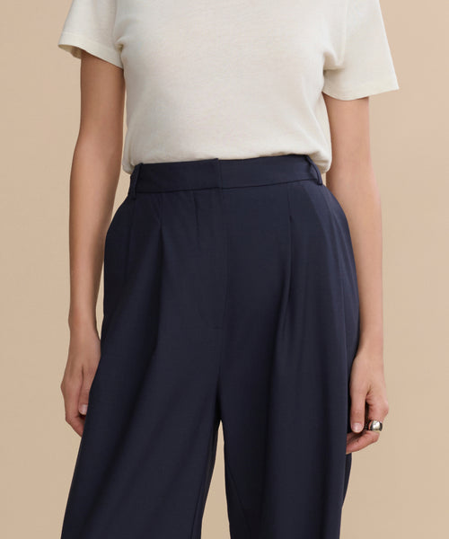 A person wears a short-sleeved, off-white t-shirt tucked into high-waisted Wool Jones Trousers in dark navy wool, standing against a plain beige background. Their left hand is visible with a ring.