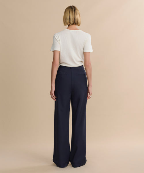 A person with short blond hair, wearing a white short-sleeve t-shirt and high-waisted, wide-leg Wool Jones Trouser in navy, stands facing away against a plain beige background.