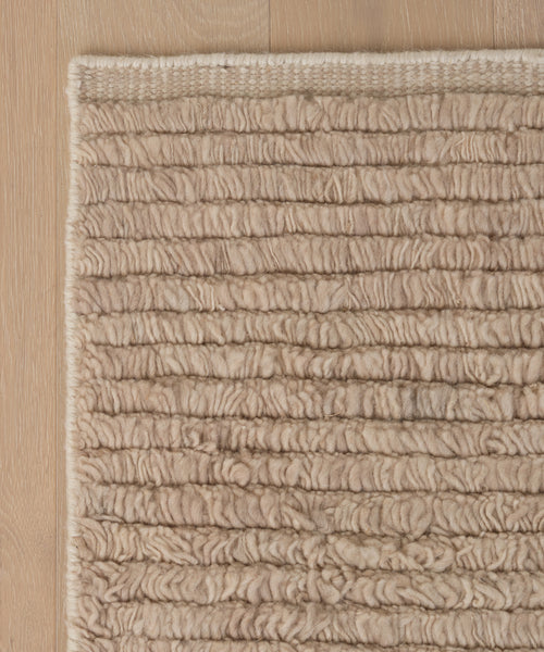 A close-up of the Winward Rug, a beige handwoven wool piece with textured, raised looped patterns, showcased on a wooden floor and highlighting its detailed woven surface.