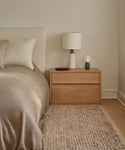 A cozy bedroom features a beige bed with neutral pillows, a wooden nightstand with a white lamp, flowers in a vase, and a decorative dish. Beside the bed, the Winward Rug adds warmth and texture to the floor.