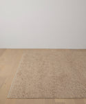 The Winward Rug, in beige handwoven wool, is placed on a light wooden floor against a plain white wall in a minimal, modern room.
