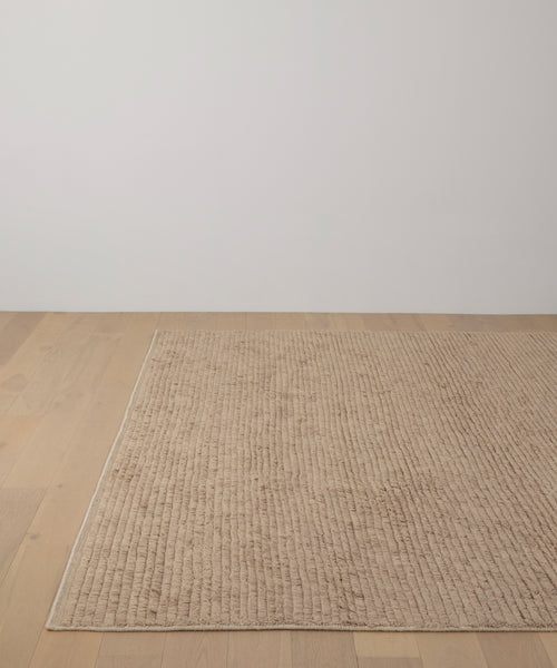 The Winward Rug, in beige handwoven wool, is placed on a light wooden floor against a plain white wall in a minimal, modern room.