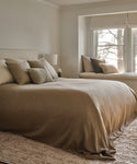 A cozy, neutral-toned bedroom features a large bed with beige bedding, plush pillows, and the Winward Rug adding warmth. A bedside table with a lamp sits nearby, while a window seat with cushions overlooks trees outside.