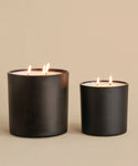 The Extra Large Topanga Glass Candle, hand-poured in Los Angeles with three flames, is lit and displayed next to a smaller two-flame black matte candle on a beige surface against a matching background.