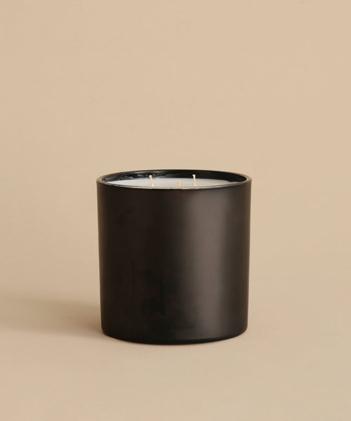 The Extra Large Topanga Glass Candle features a sleek black cylindrical design with three wicks. Hand-poured in Los Angeles, it is displayed on a beige surface with a matching beige background.