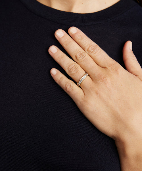 A person wearing a dark shirt rests their left hand on their chest, showcasing the Stevie Grey Diamond Band with small stones on their ring finger—a perfect piece of fine jewelry.
