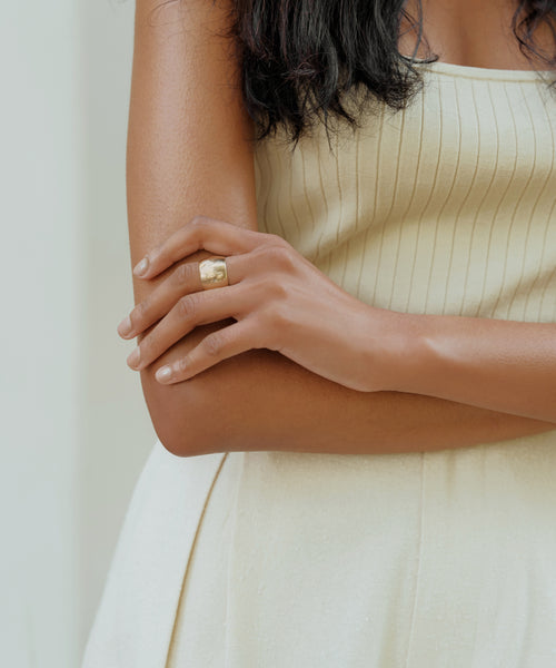 A woman in a sleeveless cream ribbed dress crosses her arms, showcasing the Campbell Cigar Band ring on her left hand. Her dark, wavy hair frames her upper body against a light background.