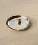 The Campbell Cigar Band, a 14k solid gold ring, rests on a shiny silver plate atop a beige textured surface, with soft natural light casting gentle shadows.