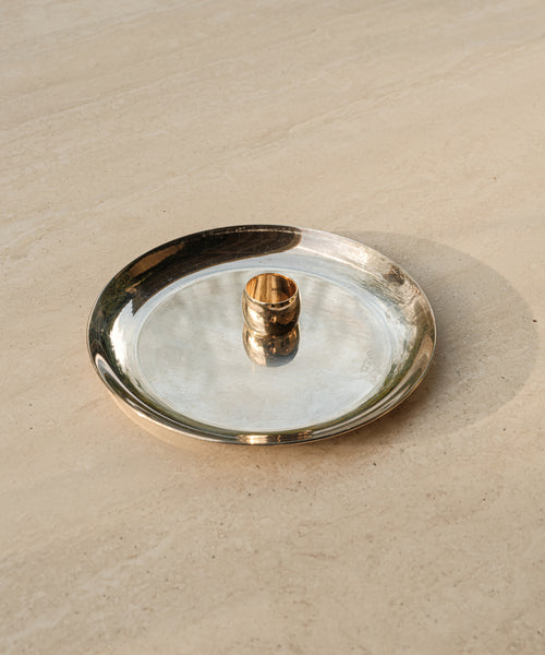 The Campbell Cigar Band, a 14k solid gold ring, rests on a shiny silver plate atop a beige textured surface, with soft natural light casting gentle shadows.
