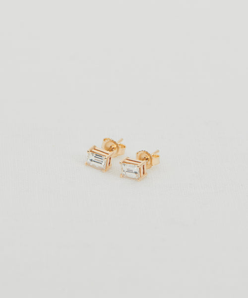 The Ivy Emerald Cut Studs are 14k gold earrings with rectangular clear gemstones in an emerald cut stud design, shown on a white textured surface.