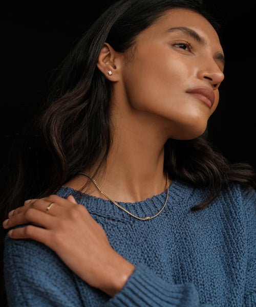 A woman wearing a blue knit sweater, gold necklaces, a ring, and Ivy Emerald Cut Studs in 14k gold looks to the side with her hand on her shoulder against a dark background.