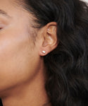 Close-up of a person’s ear wearing Ivy Emerald Cut Studs: a small, clear gemstone stud earring in 14k gold, shining against medium-brown skin and long, wavy black hair.