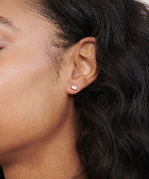 Close-up of a person’s ear wearing Ivy Emerald Cut Studs: a small, clear gemstone stud earring in 14k gold, shining against medium-brown skin and long, wavy black hair.