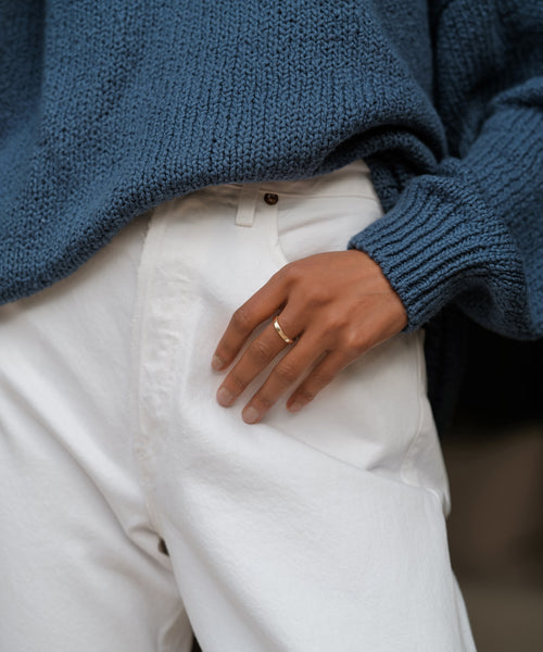 A person in a chunky blue knit sweater and white pants rests their left hand, wearing the James Band from Jenni Kayne Jewelry, in their pocket.