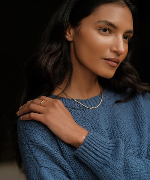 A woman with long dark hair wears a blue knit sweater and the James Band, a 14k solid gold necklace. She gently rests her hand on her shoulder, gazing calmly to the side against a dark background.