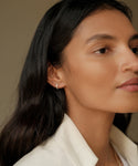 A woman with long dark hair and smooth skin wears a white blazer and Merit Baguette Hoops. She gazes calmly to the right, the timeless earrings adding elegance to her look against a neutral background.