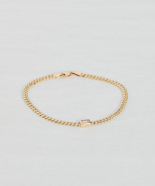 The Merit Emerald Cut Bracelet is a delicate gold piece featuring a rectangular emerald-cut gemstone centerpiece, elegantly displayed on a white fabric background—a stunning example of fine jewelry.