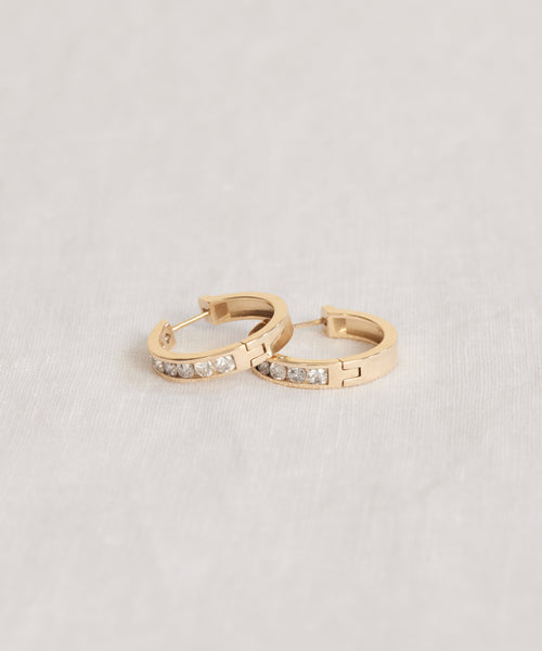 The Stevie Grey Diamond Hoops are small 14k gold earrings featuring round grey diamonds, inspired by Jenni Kayne Jewelry, shown on a neutral, light-colored fabric background.