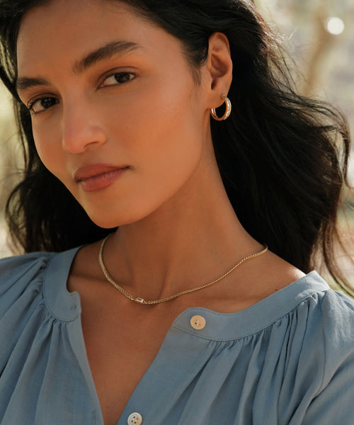 A woman with long dark hair wears a light blue blouse and Stevie Grey Diamond Hoops, paired with a delicate gold necklace, looking softly at the camera in natural outdoor light.