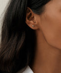 Close-up of a womans ear with long dark hair, wearing the elegant Stevie Grey Diamond Studs. The round design sparkles subtly, set in 14k gold, with a neutral background highlighting this refined Jenni Kayne jewelry piece.
