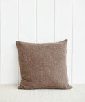 An Alpaca Basketweave Pillow in a square shape and brown color is propped upright against a white paneled wall, resting on a light-colored carpet.
