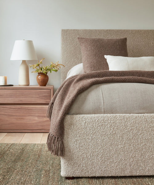 A cozy bedroom featuring a textured bed dressed in beige and taupe bedding, the Alpaca Basketweave Pillow for softness, a brown throw blanket, wooden nightstand, white lamp, floral vase, and a woven rug on the floor.