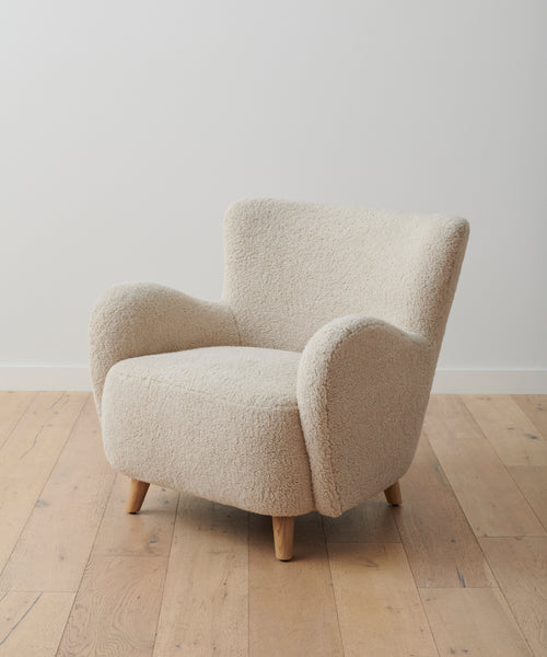 The Aspen Chair features a modern, cream-colored sherpa fabric with soft texture, rounded arms, and short angled wooden legs for a mid-century look. It sits elegantly on a wooden floor against a plain white wall.