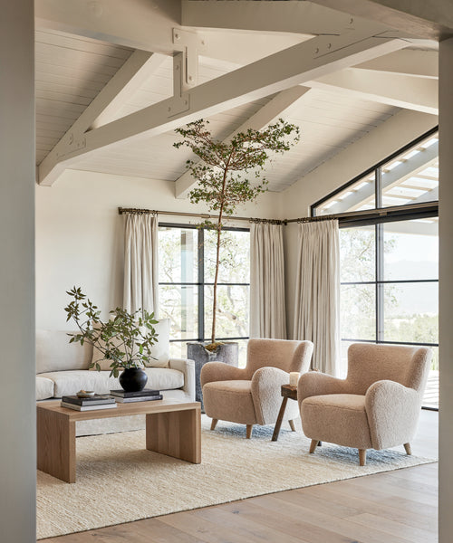 A bright, modern living room features two Aspen Chairs and a light Aspen Sofa upholstered in Fabric Swatch Oatmeal Sherpa, a wooden coffee table, neutral rug, large curtained windows, and potted plants for a fresh, minimalist vibe.