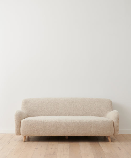 The Aspen Sofa, a simple beige mid-century modern piece with wooden legs, sits against a plain white wall on a light wood floor in a minimalistic modern room.