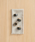 A light gray napkin is neatly folded on a wooden surface, topped with the Ceramic Napkin Rings Set of 4—each round and handmade, featuring black glaze and natural wood accents.