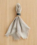 A beige fabric napkin with frayed edges is gathered through a Ceramic Napkin Rings Set of 4, handmade with a black glaze, and placed on a light wood surface.