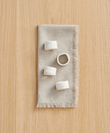 A beige linen napkin with fringe sits on wood, displaying the Ceramic Napkin Rings Set of 4—handmade in California, three upright and one on its side—all finished with an elegant ivory glaze.