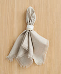 A beige linen napkin with frayed edges is folded and held by a Ceramic Napkin Rings Set of 4, handmade in California, and placed on a light wooden surface.
