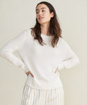 A woman with long dark hair models the Everyday Sweater in ivory, paired with cream pants featuring thin vertical stripes, standing hands on hips against a plain light background.
