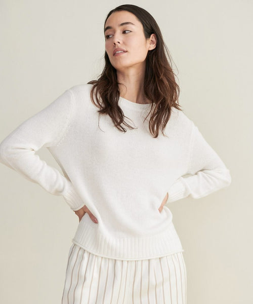 A woman with long dark hair models the Everyday Sweater in ivory, paired with cream pants featuring thin vertical stripes, standing hands on hips against a plain light background.