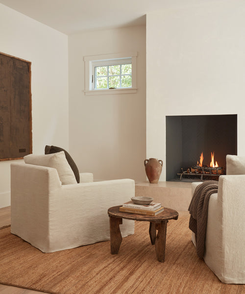 A cozy living room featuring two Harbor Chairs in natural linen, a wooden coffee table with books, a brown rug, a fireplace with a warm fire, a ceramic vase, and natural light streaming through a small window.