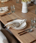 A wooden dining table set with white plates, a Ceramic Napkin Rings Set of 4 handmade in California holding beige cloth napkins, water glasses, brown-handled cutlery, and a white flower vase at the center creates a neat and inviting scene.