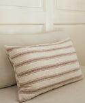 The Mill Pillow, a rectangular beige decorative pillow with brown horizontal stripes and a plush feather down insert, rests on a light-colored sofa against a cream paneled wall.