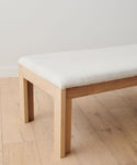 The Otto Bench, made from handmade white oak and topped with a natural linen cushion, sits on a light wood floor by a plain white wall. The image shows only one end of the bench.