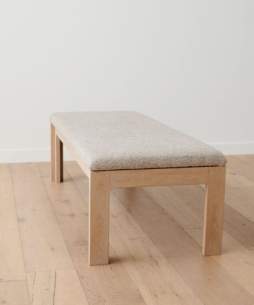 The Otto Bench, crafted in white oak with a light beige wool boucle seat, rests on a light wood floor against a plain white wall.