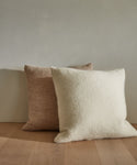 A cream Alpaca Basketweave Pillow sits on a light wooden floor in front of a larger beige pillow, both resting against a plain gray wall.