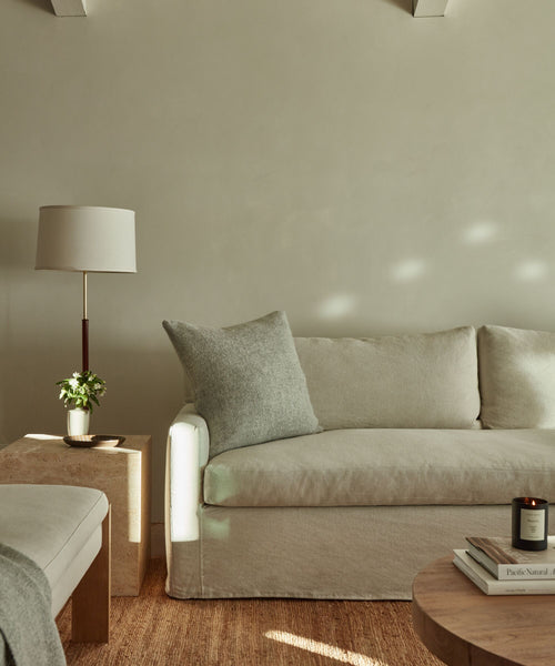 A minimalist living room with a light beige sofa, an Alpaca Basketweave Pillow, a wooden side table holding a lamp and small plant, a round coffee table with books and a candle, and neutral-toned walls.