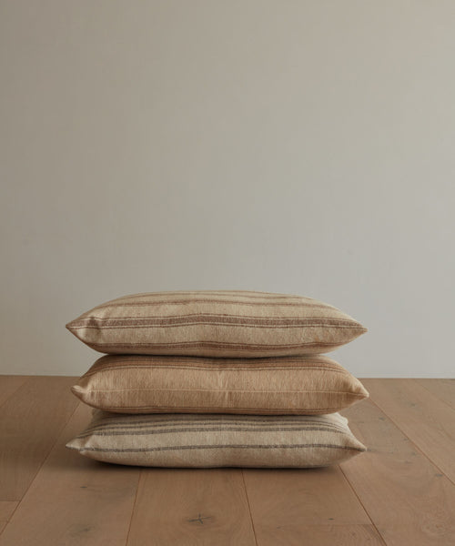 Three Mill Pillows with beige and brown stripes are stacked on a wooden floor against a plain, light wall, each featuring a plush feather down insert for added comfort.