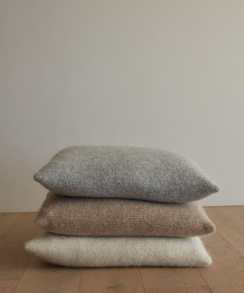 Three textured, knitted throw pillows—including the super-soft Alpaca Basketweave Pillow in gray, beige, and off-white—are stacked on a wooden floor against a plain light-colored wall.