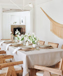Bright dining room with a wooden table set for six, featuring the Frayed Linen Tablecloth, neutral plates, napkins, wicker placemats, glassware, and white flower centerpieces; cozy rustic style enhanced by wooden chairs and a fringed wall hanging.