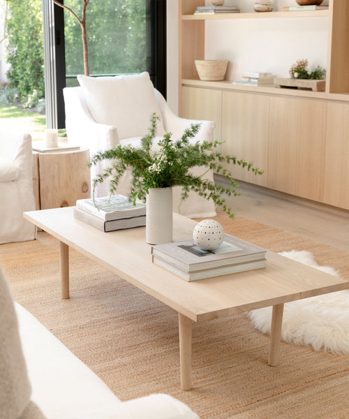 The Arrowhead Jute Rug anchors a bright, minimalist living room featuring a light wood coffee table with books, a white vase of greenery, and decor, surrounded by neutral furniture, a window, and built-in shelves.
