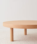 The Bay Coffee Table, crafted from knotty alder, showcases a minimalist round form with three cylindrical legs and is displayed on a light textured carpet in front of a plain white wall.