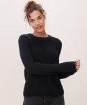 A woman with curly hair in a loose bun wears the Cotton Fisherman Sweater and dark pants, standing against a plain light background and looking at the camera with a slight smile.