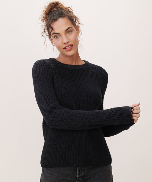 A woman with curly hair in a loose bun wears the Cotton Fisherman Sweater and dark pants, standing against a plain light background and looking at the camera with a slight smile.